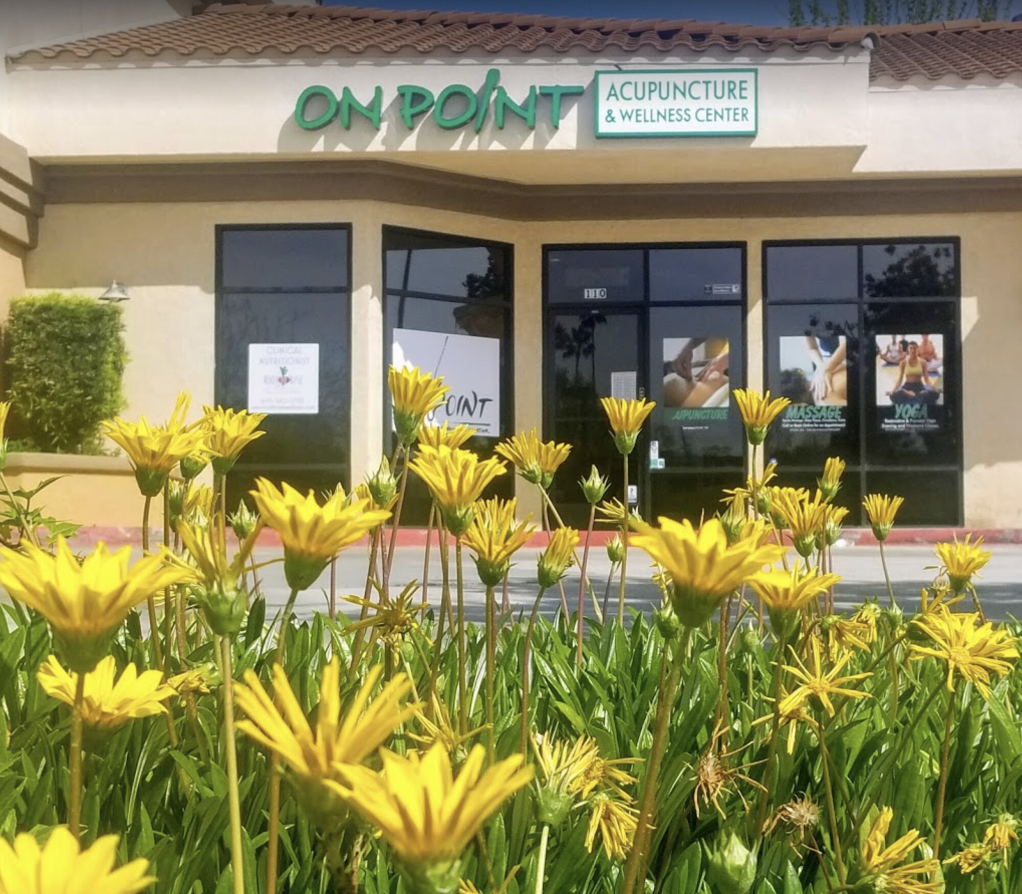 Exterior storefront of On Point with green signage behind a bed of yellow flowers